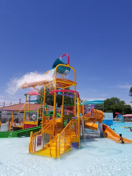 Colorful water slide structure with splashes, surrounded by a pool and sunny sky, with children playing nearby.