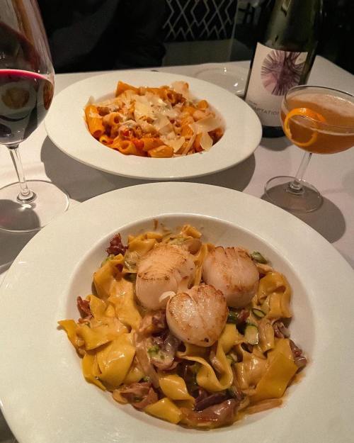 A table setting with two pasta dishes, one with scallops, a glass of red wine, and a cocktail.
