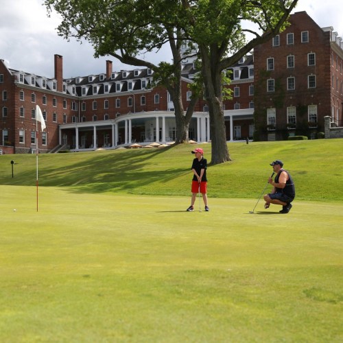 A child watches as an adult prepares to putt on a golf course, with a historic building in the background.