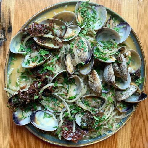 A plate of clams on a bed of noodles, garnished with herbs and seaweed, served in a flavorful broth.