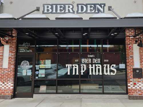 Exterior of Bier Den Tap Haus, featuring large glass windows and a brick facade, with the establishment's name prominently displayed.