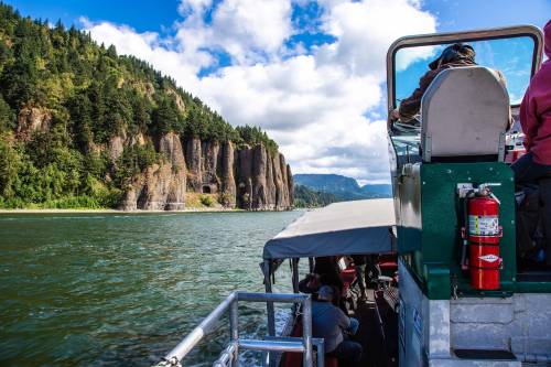 A boat on a river with rocky cliffs and lush greenery under a partly cloudy sky. Passengers enjoy the scenic view.