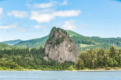 A tall, rocky outcrop surrounded by lush green trees and mountains, with a calm river in the foreground under a blue sky.