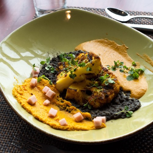 A plate featuring grilled pineapple, black bean puree, orange sauce, and garnished with herbs and diced ham.