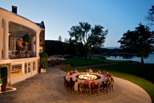 A group of people gathered around a fire pit at dusk, with a scenic view of a lake and a building in the background.