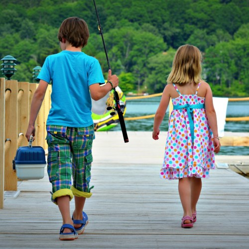 Two children walk along a wooden dock, one carrying a fishing rod and the other a small cooler, surrounded by nature.