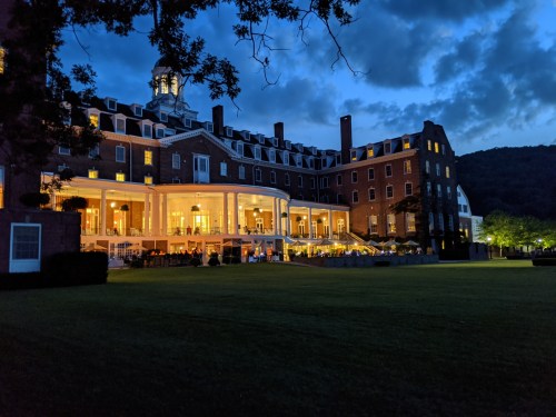 A historic hotel illuminated at dusk, surrounded by trees and mountains, with outdoor dining visible on the patio.
