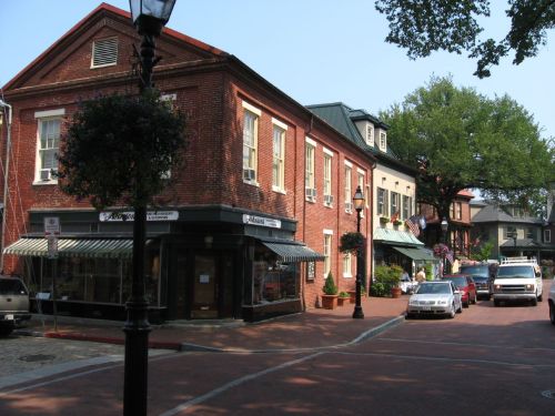 Charming brick buildings line a quiet street, with parked cars and greenery adding to the quaint atmosphere.