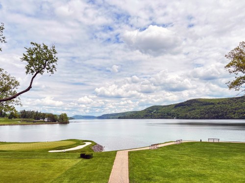 A serene lake view with green grass, a pathway, and rolling hills under a cloudy sky.