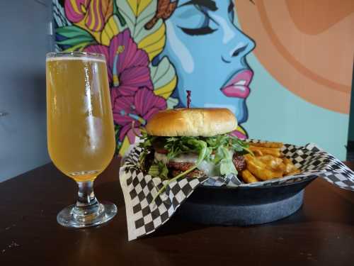 A burger with lettuce and tomato, served with fries, alongside a glass of beer, against a colorful mural backdrop.