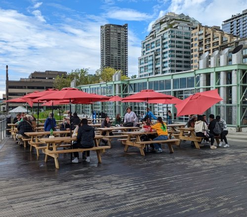 Outdoor seating area with picnic tables and red umbrellas, filled with people enjoying a sunny day in an urban setting.