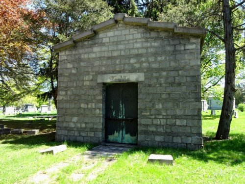A small, stone mausoleum with a green door, surrounded by grass and trees in a cemetery.