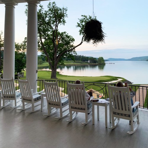 A serene lakeside view from a porch with rocking chairs, featuring people enjoying drinks and the peaceful landscape.