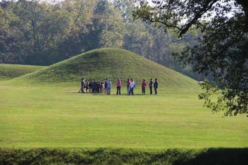 A group of people gathers near a large grassy mound in a park, surrounded by trees and open green space.