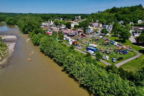 Aerial view of a riverside park with people, tents, and activities, surrounded by greenery and a small town in the background.