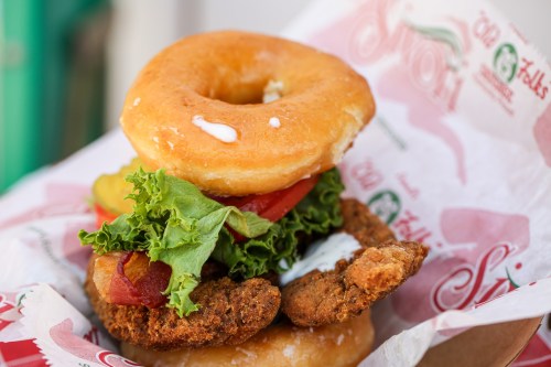 A stacked sandwich featuring a glazed donut, fried chicken, lettuce, tomato, and pickles, served in a paper wrapper.