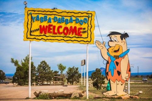A colorful sign reading "Yabba Dabba Doo Welcome" with a cartoon character waving, set against a blue sky.