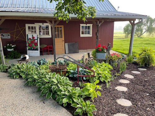 A charming red barn with a garden featuring hostas, flowers, and decorative items, set in a serene landscape.