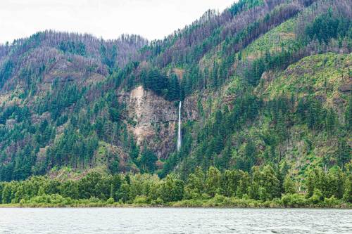 A scenic view of a waterfall cascading down a green mountain, surrounded by trees and a calm river in the foreground.
