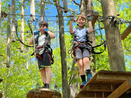 Two children smiling on a ropes course, wearing harnesses and gloves, surrounded by trees and blue sky.