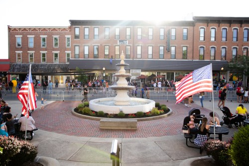 A bustling town square with a fountain, American flags, and people enjoying the outdoor space on a sunny day.