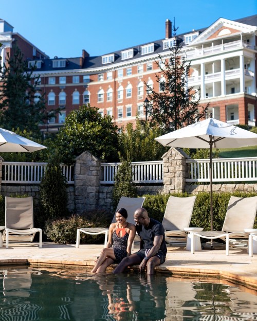 A couple sits by a pool, dipping their feet in the water, with a historic building and trees in the background.