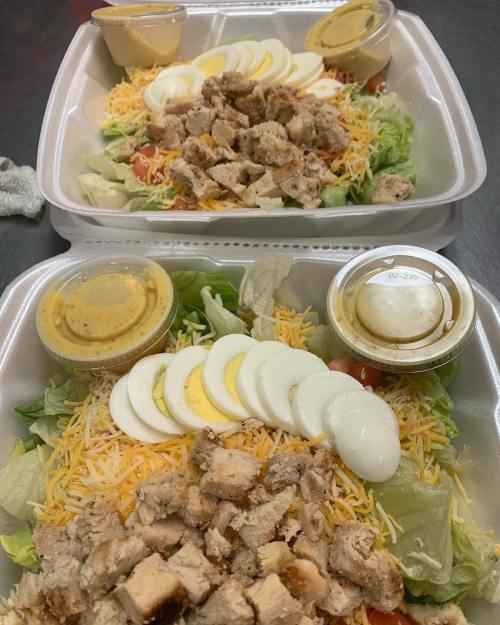 Two salad containers with chopped chicken, shredded cheese, sliced eggs, lettuce, tomatoes, and dressing on the side.