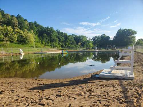 A calm pond surrounded by trees, with a sandy beach and a lifeguard stand in the foreground.