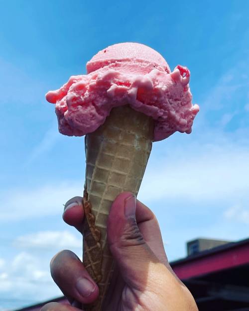 A hand holding a cone of pink ice cream against a bright blue sky.