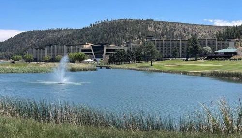 A serene lake with a fountain, surrounded by lush greenery and a hotel in the background against a mountainous landscape.