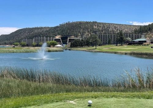 A scenic view of a golf course with a lake, fountain, and mountains in the background under a clear blue sky.