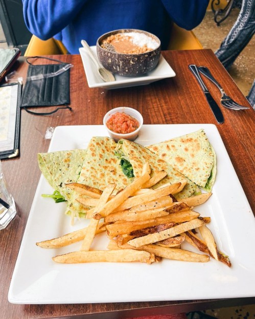 A plate with green quesadillas, fries, and salsa, with a bowl of soup in the background on a wooden table.