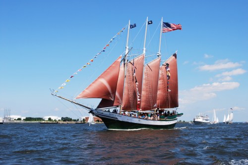 A tall ship with brown sails and flags sails on blue water under a clear sky, with people on board enjoying the view.
