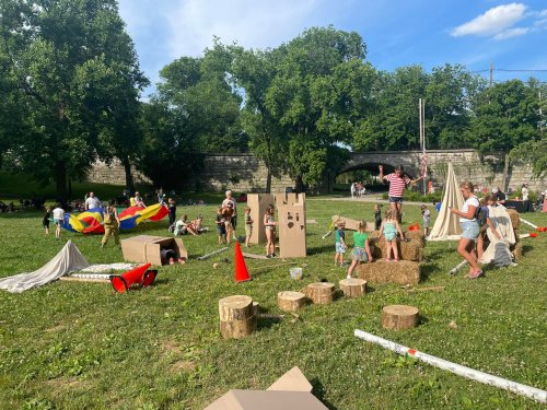 Children play in a grassy park with cardboard structures, tents, and colorful parachutes under a blue sky.