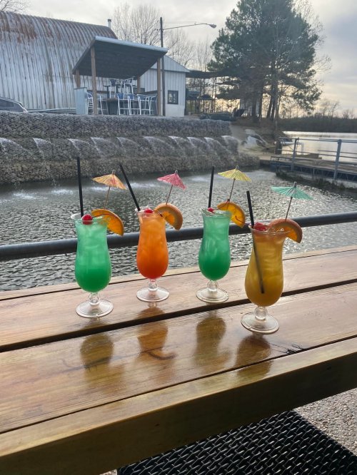 Four colorful cocktails with umbrellas and fruit garnishes sit on a wooden table by a scenic waterfront.