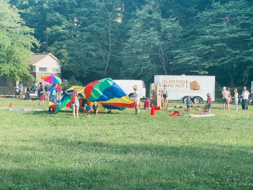 A colorful parachute activity in a park with people gathered around, surrounded by trees and a food truck in the background.