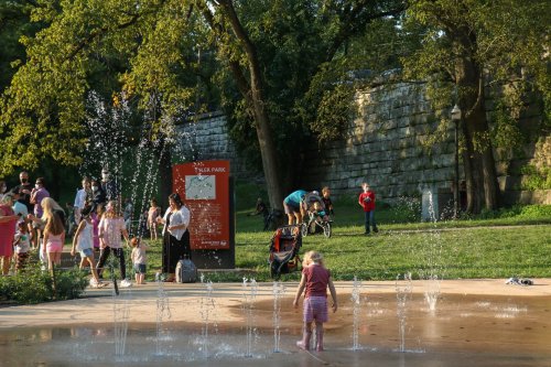 A child plays in a splash pad while families enjoy the park on a sunny day, with a stone wall in the background.