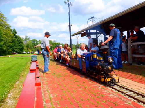 A miniature train with passengers on a track, surrounded by people and greenery on a sunny day.