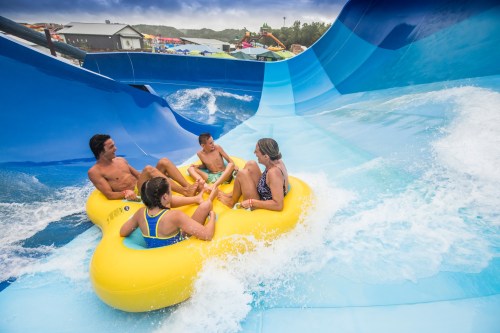 Four people in inner tubes enjoy a water slide, splashing through blue waves at a water park.
