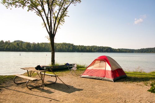 A red and gray tent by a calm lake, with a picnic table and a tree in a serene outdoor setting.