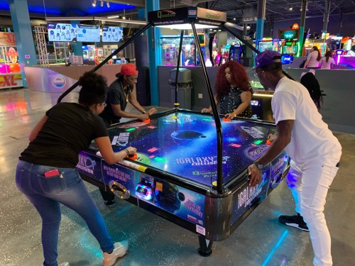 Four people playing air hockey at an arcade, with colorful lights and games in the background.
