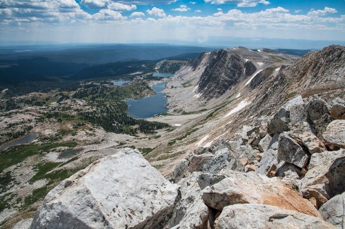 A panoramic view of rugged mountains and lakes under a blue sky with scattered clouds, showcasing rocky terrain.