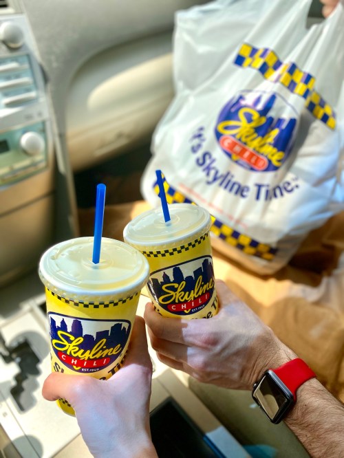 Two hands holding Skyline Chili drinks in the car, with a takeout bag in the background.