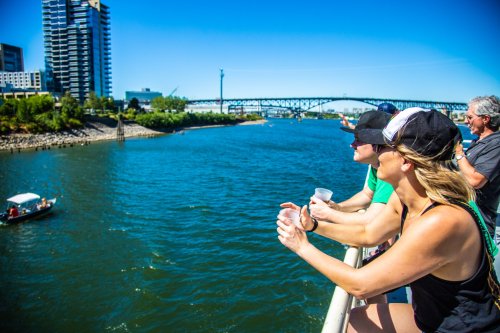 Two people lean on a railing by a river, enjoying the view of a bridge and a boat on a sunny day.