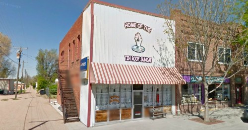 A quaint ice cream shop with a striped awning, featuring a sign that reads "Home of the Tin Roof Sundae."