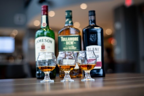 Three whiskey glasses filled with amber liquid, in front of bottles of Jameson, Tullamore Dew, and Slane whiskey.