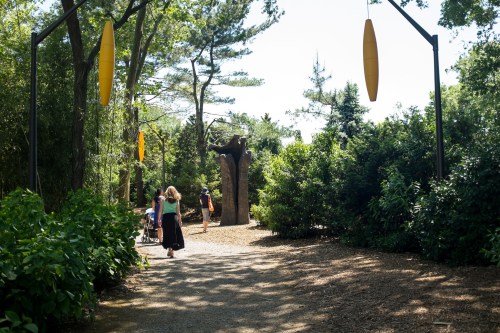 A sunny park path with visitors walking among trees and sculptures, surrounded by lush greenery.