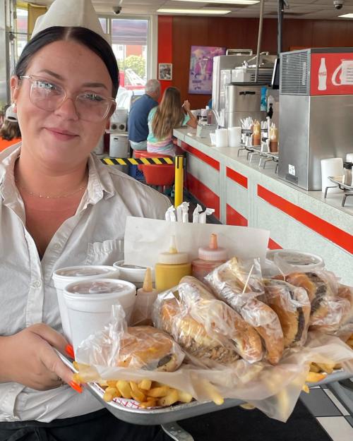 A server holds a tray with burgers, fries, and drinks in a retro diner setting. Customers are seated in the background.