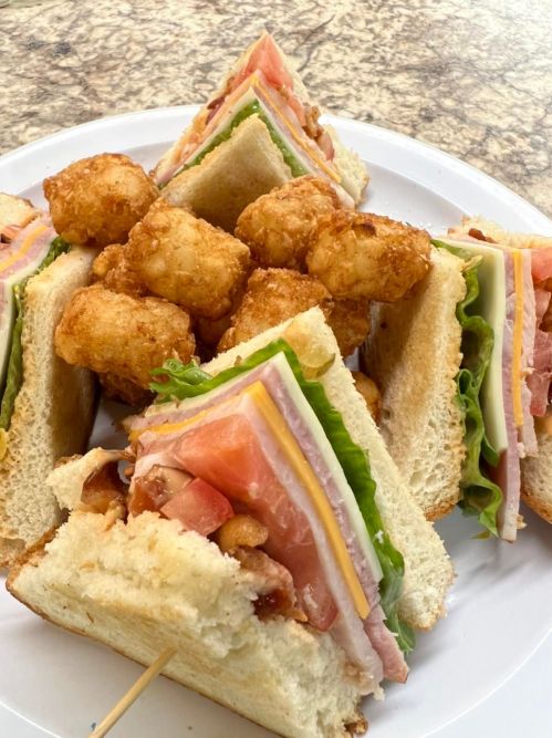 A plate with a club sandwich stacked with lettuce, tomato, ham, and cheese, accompanied by crispy tater tots.