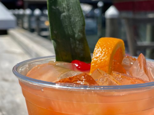 A close-up of a tropical cocktail with ice, garnished with a cherry, orange slice, and a green leaf.